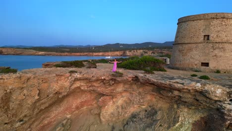 carefree woman in pink dress with open arms enjoying freedom and breathtaking view on ibiza island near torre des savinar at sunset. magic aerial view flight descending drone