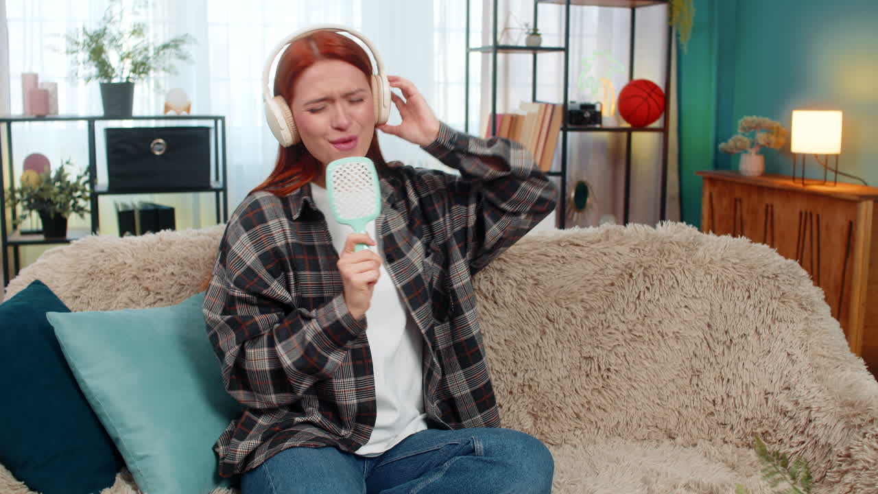 Woman sitting on sofa wearing headphones singing with comb as microphone having fun with music joy