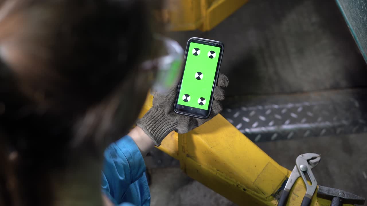 vista del hombro de una mujer mecánica de automóviles con gafas y mono sosteniendo un teléfono inteligente en la mano y mirando una pantalla verde con una llave de croma. material de video. teléfono móvil.