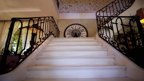 marble staircases with black wrought-iron railings, symmetrically placed green plants and an exposed stone wall with a huge clock in the background