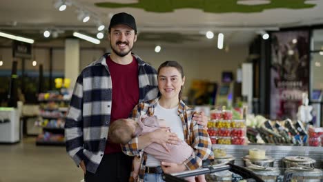 Portrait-of-a-happy-brunette-guy-with-his-wife-and-small-baby-during-family-shopping-in-a-supermarket