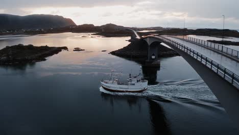 aerial view, pushing-in, tilting down, towards a fisher boat returning to port under the flakstadbruene bridge, near fredvang, lofoten islands, norway