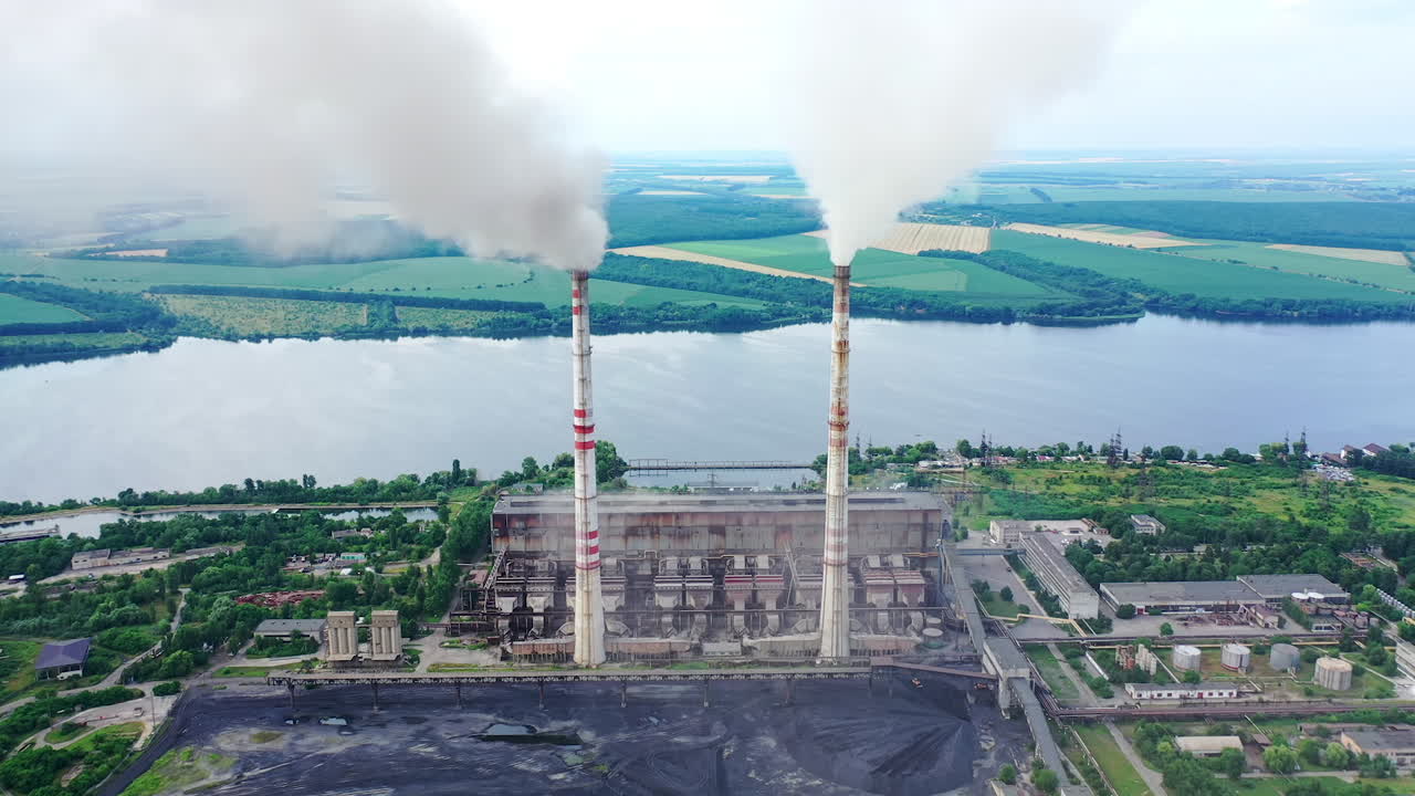 Factory with smokestacks near the river. High chimneys with thick emissions into the atmosphere on beautiful nature background. Air pollution. Aerial view.