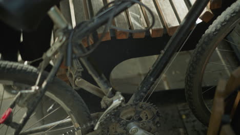 close-up of dirty bicycle being moved into place between wooden bench and nearby structure in a peaceful park, the rider lets the bike rest on the bench as he sits down on the bench