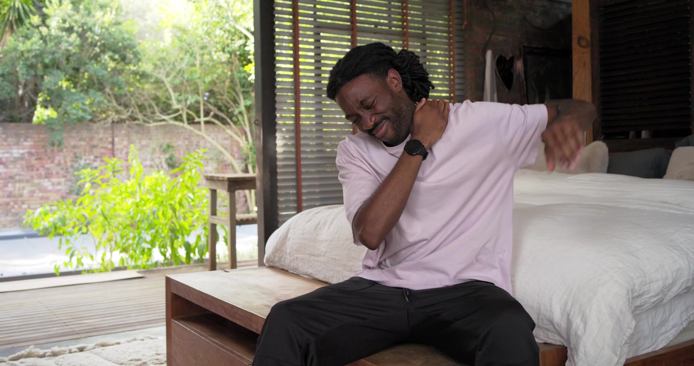 Camera settling, man sitting on wooden bench in bedroom by deck massaging neck relieving tension