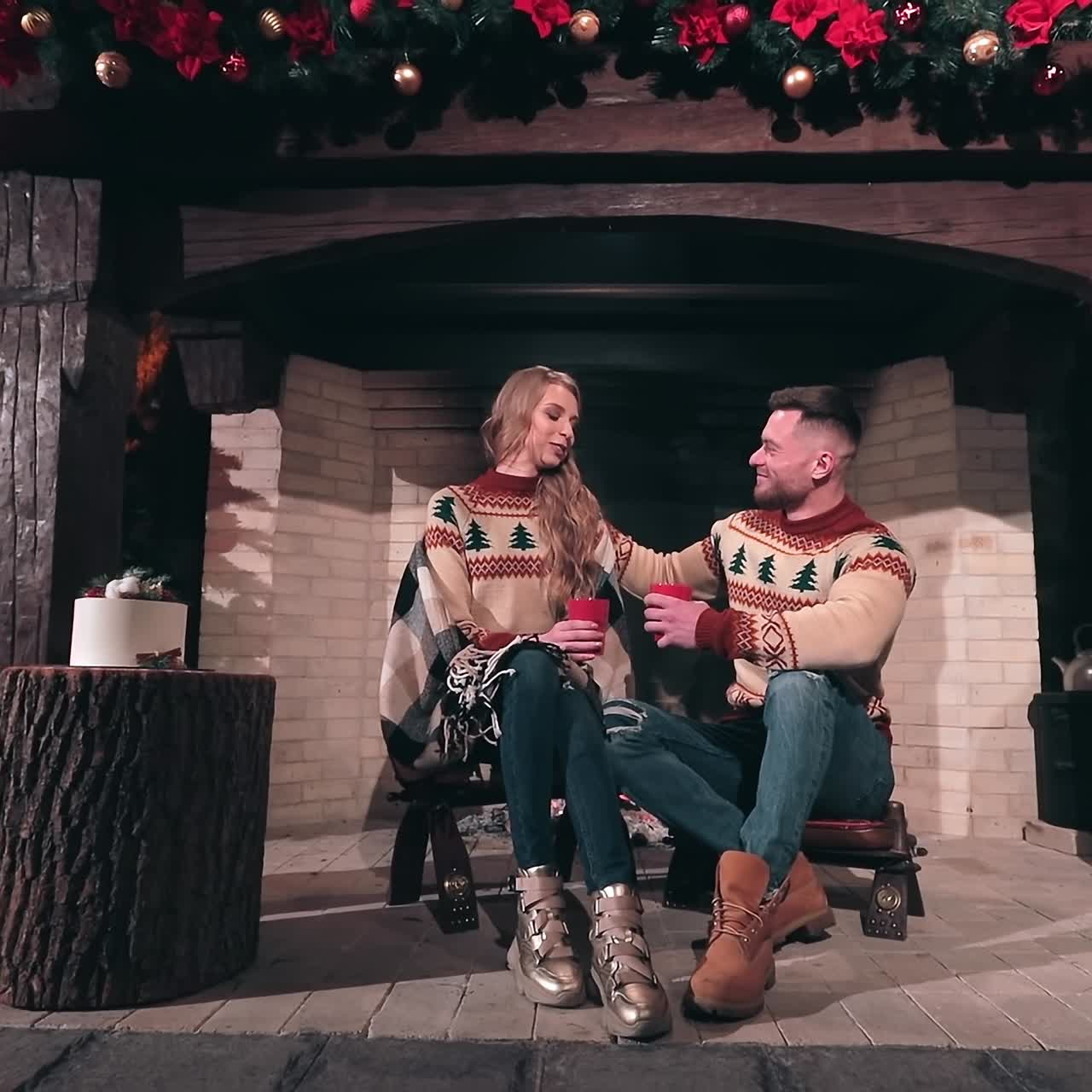 Couple in love sitting at fireplace. Young romantic pair in knitted Christmas sweaters sitting together and drinking tea. Christmas atmosphere