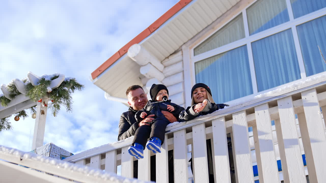Caucasian family with little toddler son stand near the house outdoors. Lovely kid sits on fence supported by dad and mom makes a snowball and throws to camera. Low angle view.