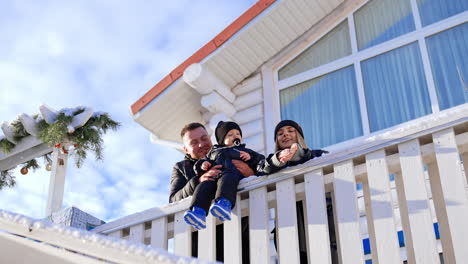 caucasian family with little toddler son stand near the house outdoors. lovely kid sits on fence supported by dad and mom makes a snowball and throws to camera. low angle view.