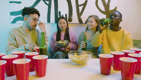 happy multiethnic young friends drinking beer, eating potato chips and talking while sitting on sofa at home