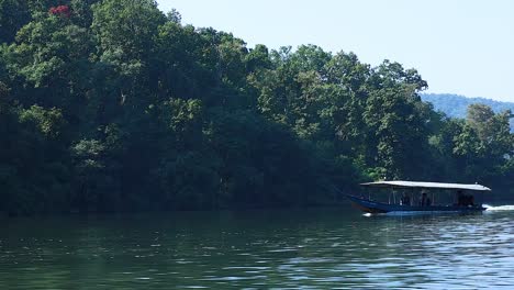A covered boat travels smoothly beside a dense, green forest along a tranquil waterway.