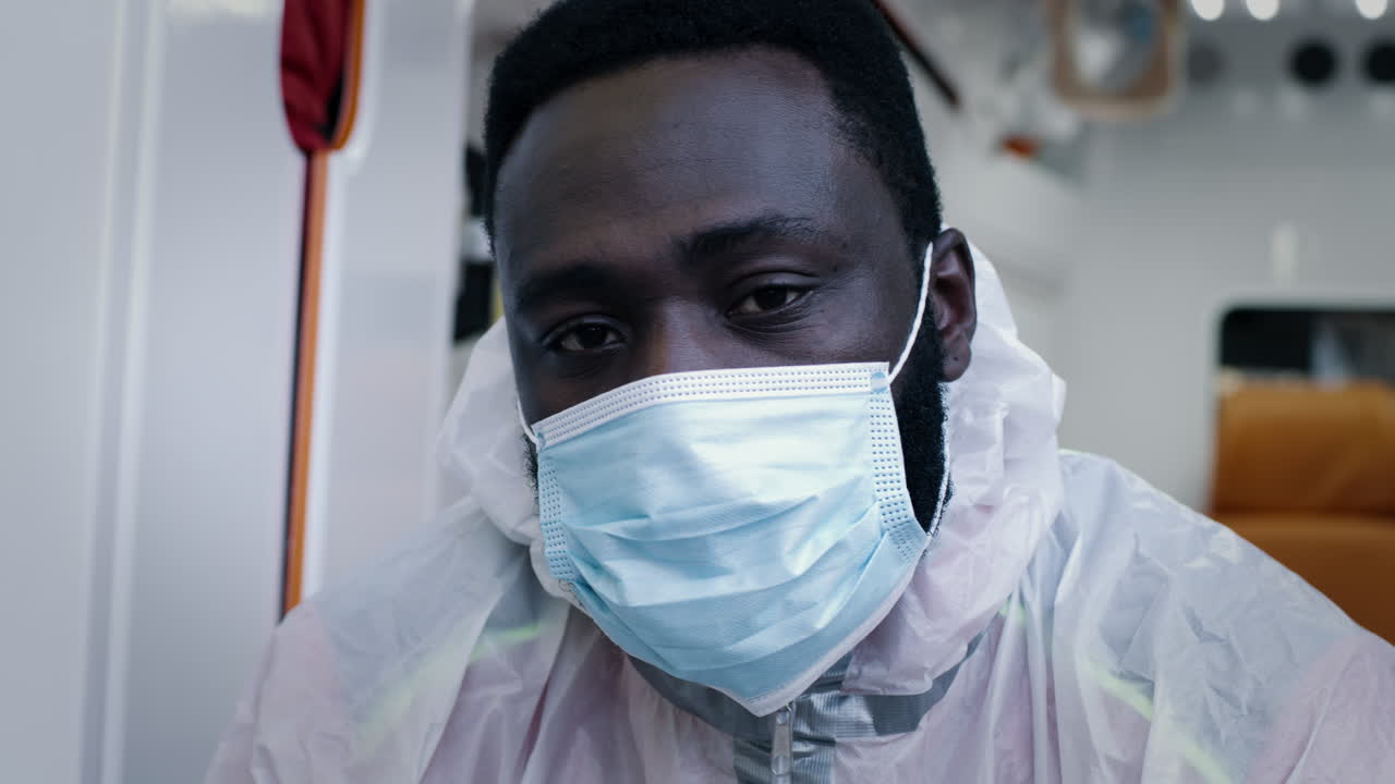 black Emergency Service Worker Taking off Face Shield African American Paramedic in Mask Taking off Face Shield and Hazmat Suit Hood while Looking at Camera in Ambulance during Pandemic