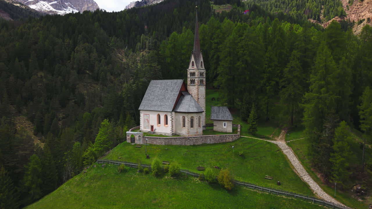 Drone footage of a historic alpine church surrounded by dense evergreen forest and mountain peaks in the Dolomites, Italy