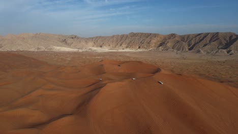 Wüste-Der-Vereinigten-Arabischen-Emirate:-Blick-Von-Oben-Auf-Die-Wüste-Von-Sharjah,-Eine-Gruppe-Von-4x4-Fahrzeugen,-Fahrten-Auf-Riesigen-Sanddünen,-Mleiha-Berge-Im-Hintergrund