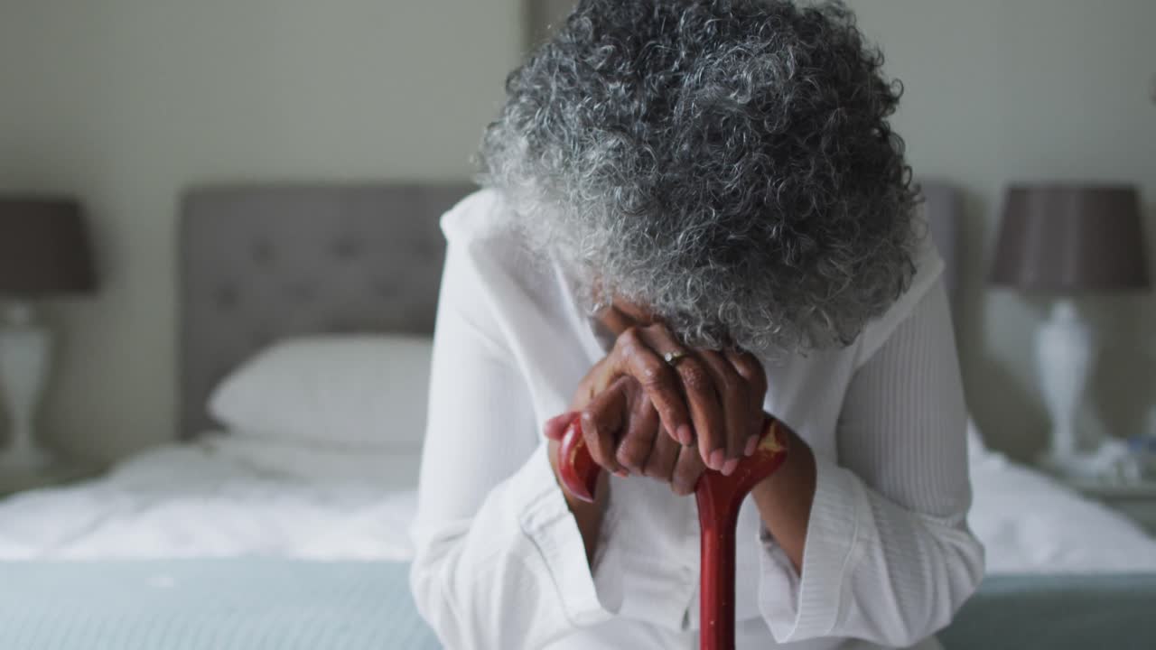 Oudere Afro-Amerikaanse vrouw die thuis op bed zit en een wandelstok vasthoudt.