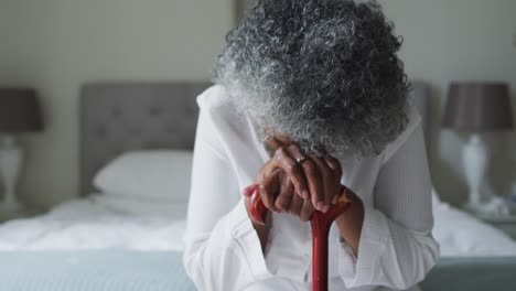 Senior-african-american-woman-holding-walking-stick-sitting-on-the-bed-at-home