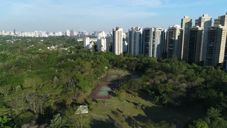 Drone-footage-with-moving-forward-showing-beautiful-tennis-courts-in-the-middle-of-a-public-park-with-lush-nature-and-buildings-in-the-background,-sport,-leisure,-health,-in-4-resolution