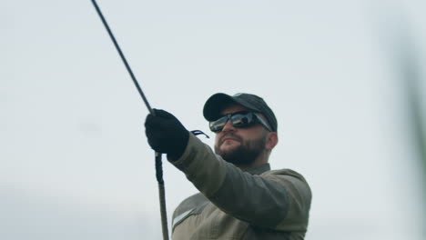 man in dark shades and cap gradually releasing tether rope connected to hot air balloon, standing amid tall grass under pale evening sky
