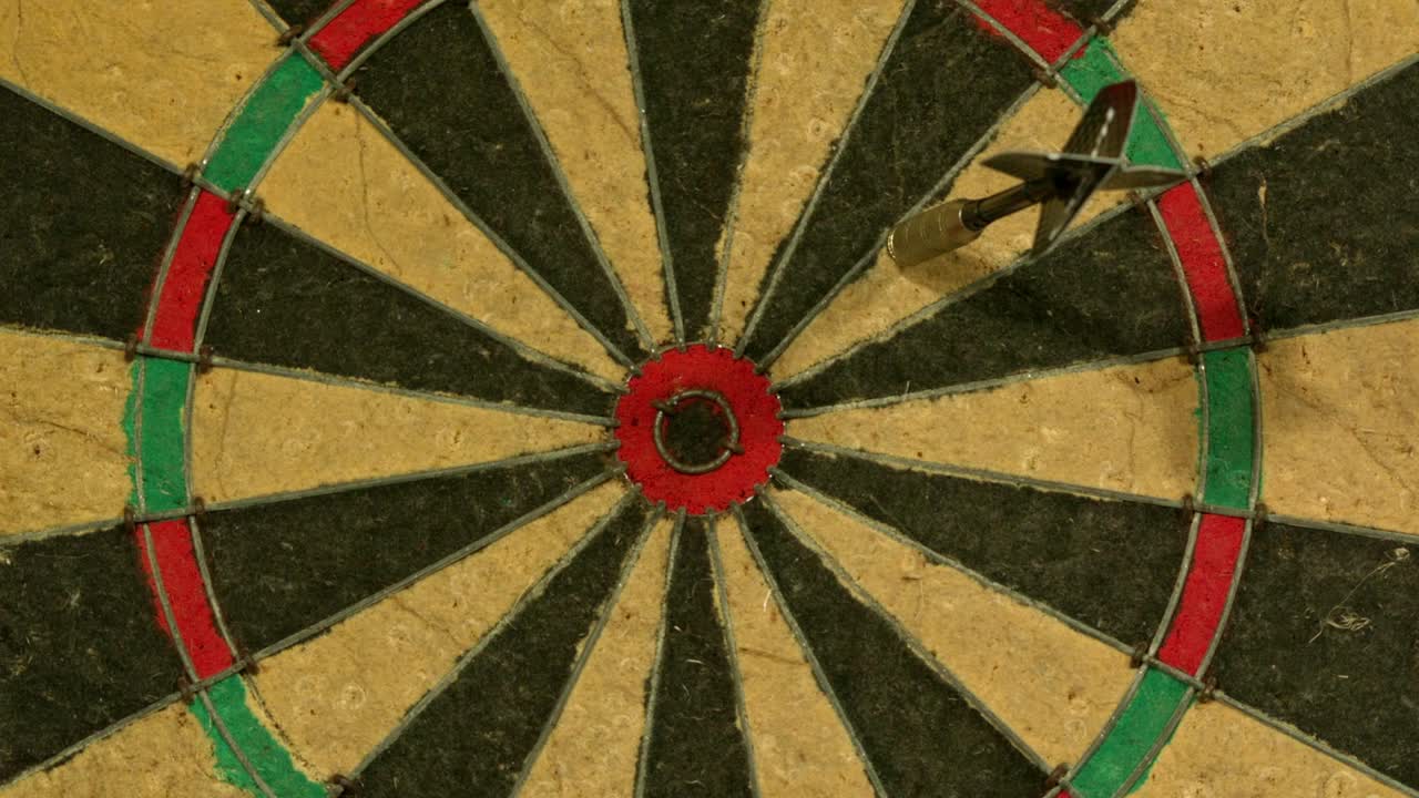Imagens em câmera lenta de um dardo atingindo um alvo de dardos tradicional, caindo no anel de pontuação vermelho logo fora do centro do alvo. O close-up captura a precisão e o impacto do arremesso