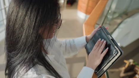 overhead view of young woman using touchscreen tablet with long manicured nails, standing by wooden railing inside sunlit glass building with smooth reflections and clean architectural lines