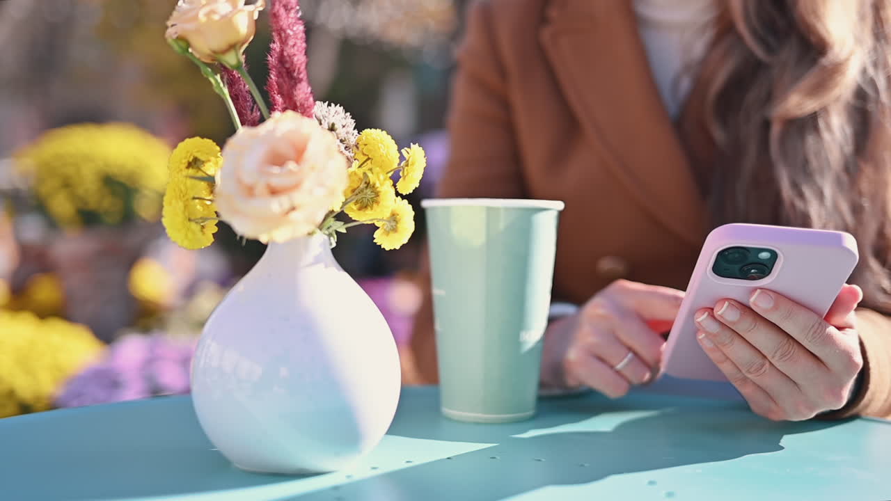 Woman in brown coat scrolling on her phone near a cup of tea at a terrace