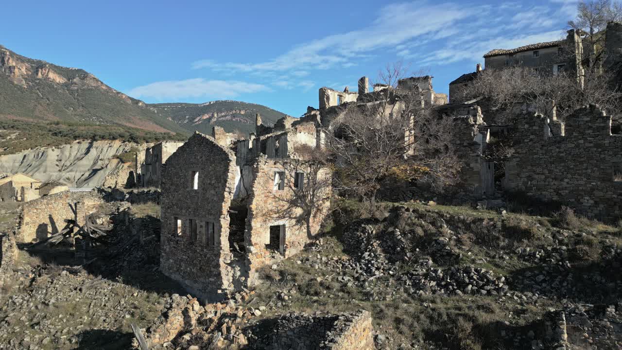 Vista aerea che circonda la città fantasma desolata e abbandonata di Esco in Aragona, Spagna, rivelando edifici in pietra fatiscenti e il paesaggio montuoso circostante sotto un cielo azzurro e limpido