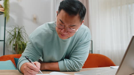 asian man freelancer making notes on notebook at home, use laptop, prepare business financial report