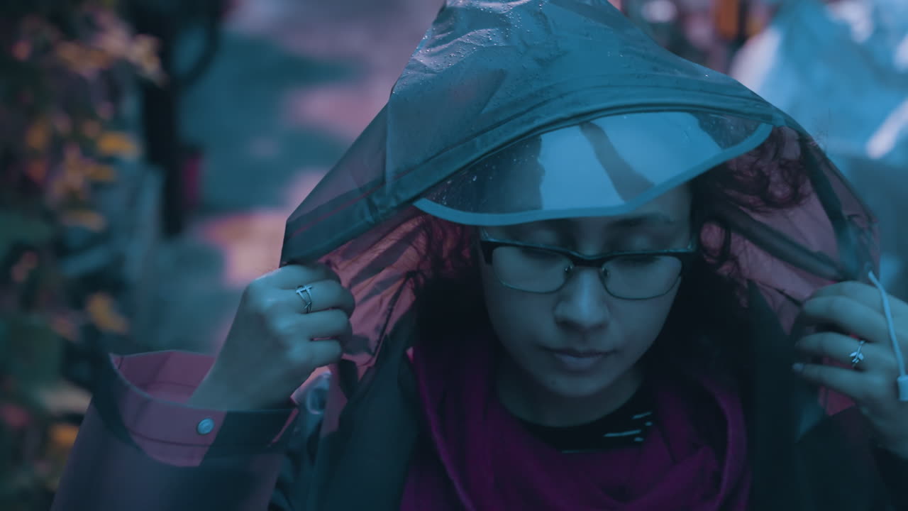 Giovane donna che cammina in un vicolo piovoso, aggiustando cappuccio e occhiali mentre indossa giacca e zaino, luce tenue al neon e del lampione, pozzanghere sulla pavimentazione, atmosfera urbana tranquilla con un umore pensieroso e solitario.
