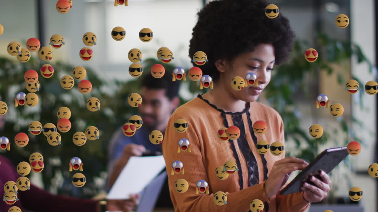 animación de iconos emoji digitales sobre una mujer de negocios biracial sonriente usando una tableta
