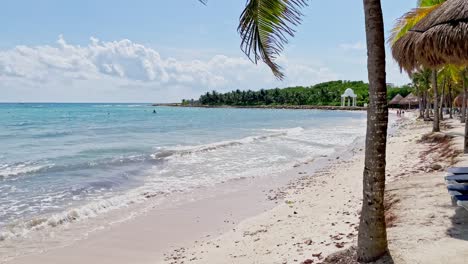 Toma-Panorámica-Lenta-A-La-Izquierda-En-Una-Hermosa-Playa-En-Tulum,-Cerca-De-Cancún,-México,-Con-Pequeñas-Olas,-Palmeras,-Cielo-Azul-Y-Algunas-Nubes.