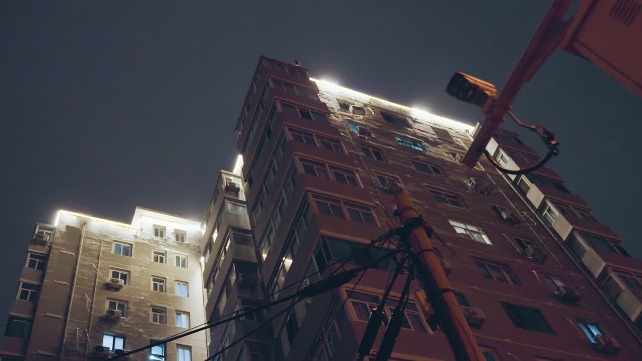 Cielo notturno di un grattacielo illuminato sul tetto, luci a stringa lungo il profilo del tetto e finestre illuminate, cavi elettrici e antenna in silhouette contro il cielo nuvoloso, grana cinematografica malinconica e prospettiva verticale, bagliore caldo.