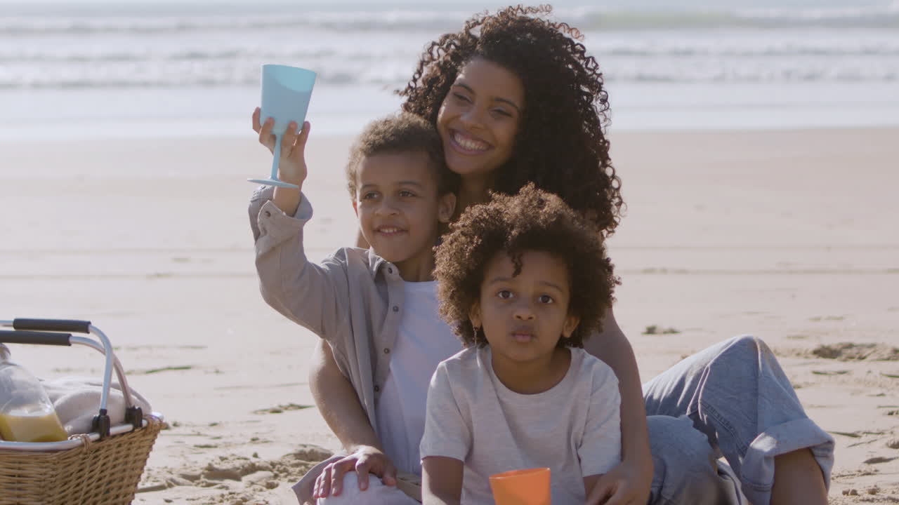 mujer negra sonriente y dos niños adorables haciendo un picnic a la orilla del mar en un día soleado
