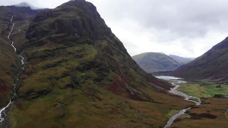 Espectacular-Toma-Aérea-Hacia-Atrás-De-Las-Montañas-Glencoe-Escocia-Que-Muestra-Un-Río-Que-Fluye-Por-La-Ladera-De-La-Montaña