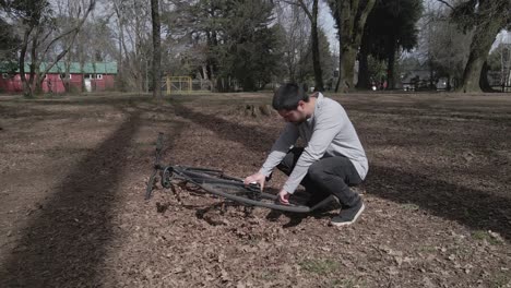 short haired man fixing broken down bike in anopen park with some trees in the background