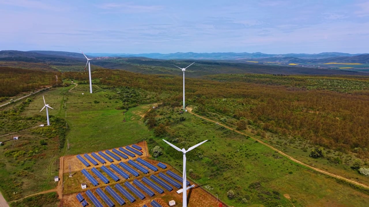 Vista aérea de gran angular de una instalación de turbina eólica y paneles solares. Generador blanco entre módulos fotovoltaicos azules sobre suelo marrón, perspectiva cenital