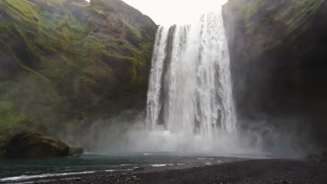 majestic waterfall cascading down mossy cliffs into a serene pool with mist rising above the black pebble beach in iceland