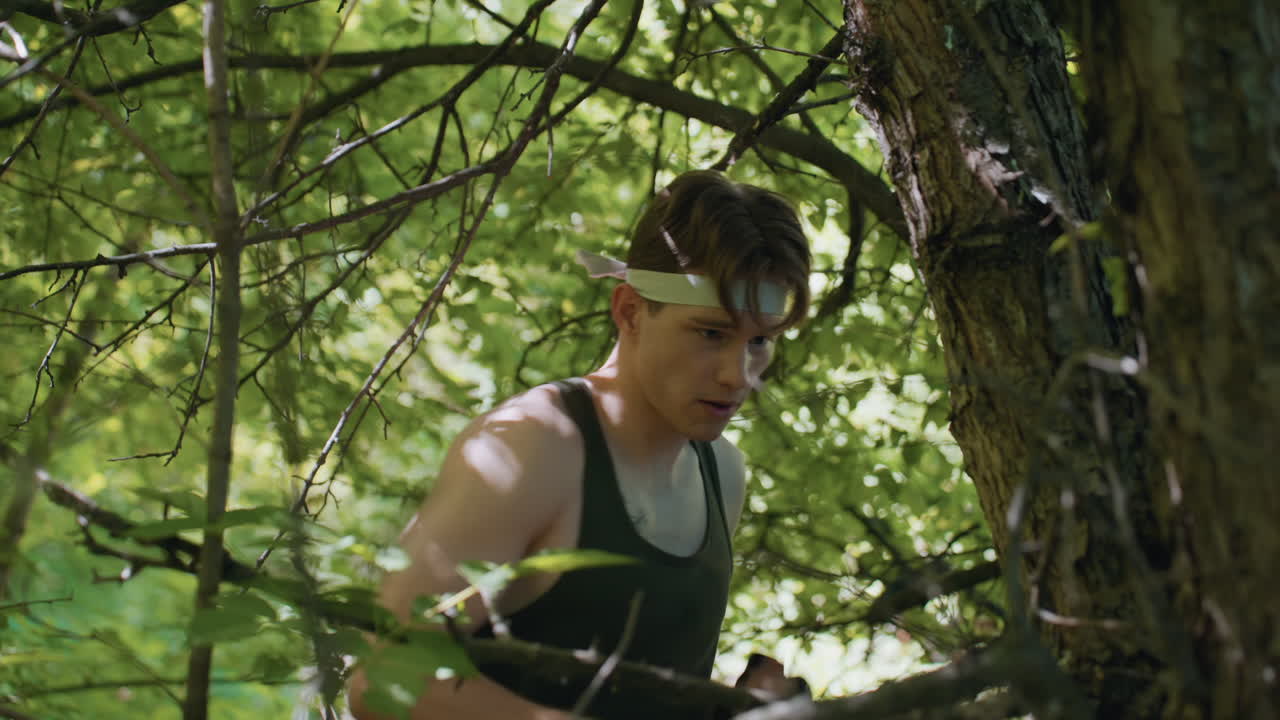 soldier wearing gloves and headband punches tree bark in dense woodland, exhales sharply while training combat technique amid tangled branches and sunlit foliage, demonstrating strength and focus