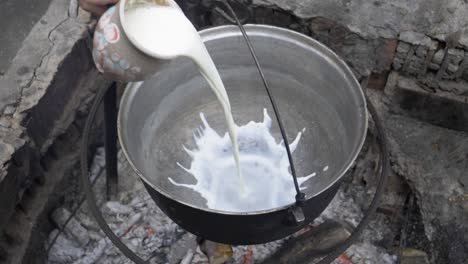 pour creamy milk into a black cauldron and boil over high heat. the location is in nature in a dense forest near a cottage
