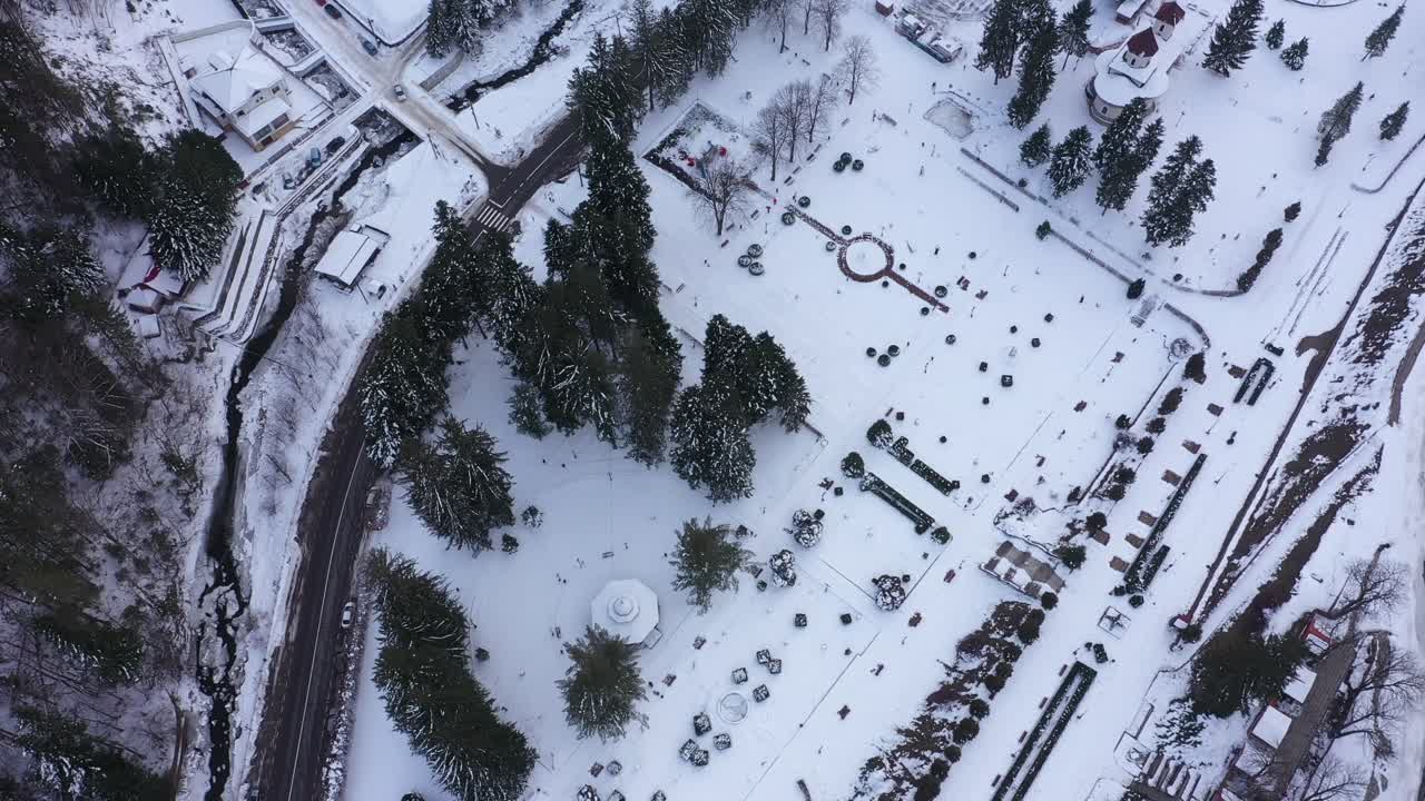 Ascending aerial view, drone, of Slanic Moldova's central park, Romania, covered in white snow. A road, river, and buildings surround the idyllic winter landscape