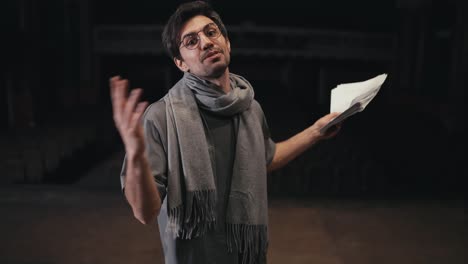 Portrait-of-a-confident-young-brunette-man-in-glasses-of-a-confident-stage-director-in-a-gray-T-shirt-and-a-gray-scarf-with-a-script-in-his-hands-on-stage-in-the-theater