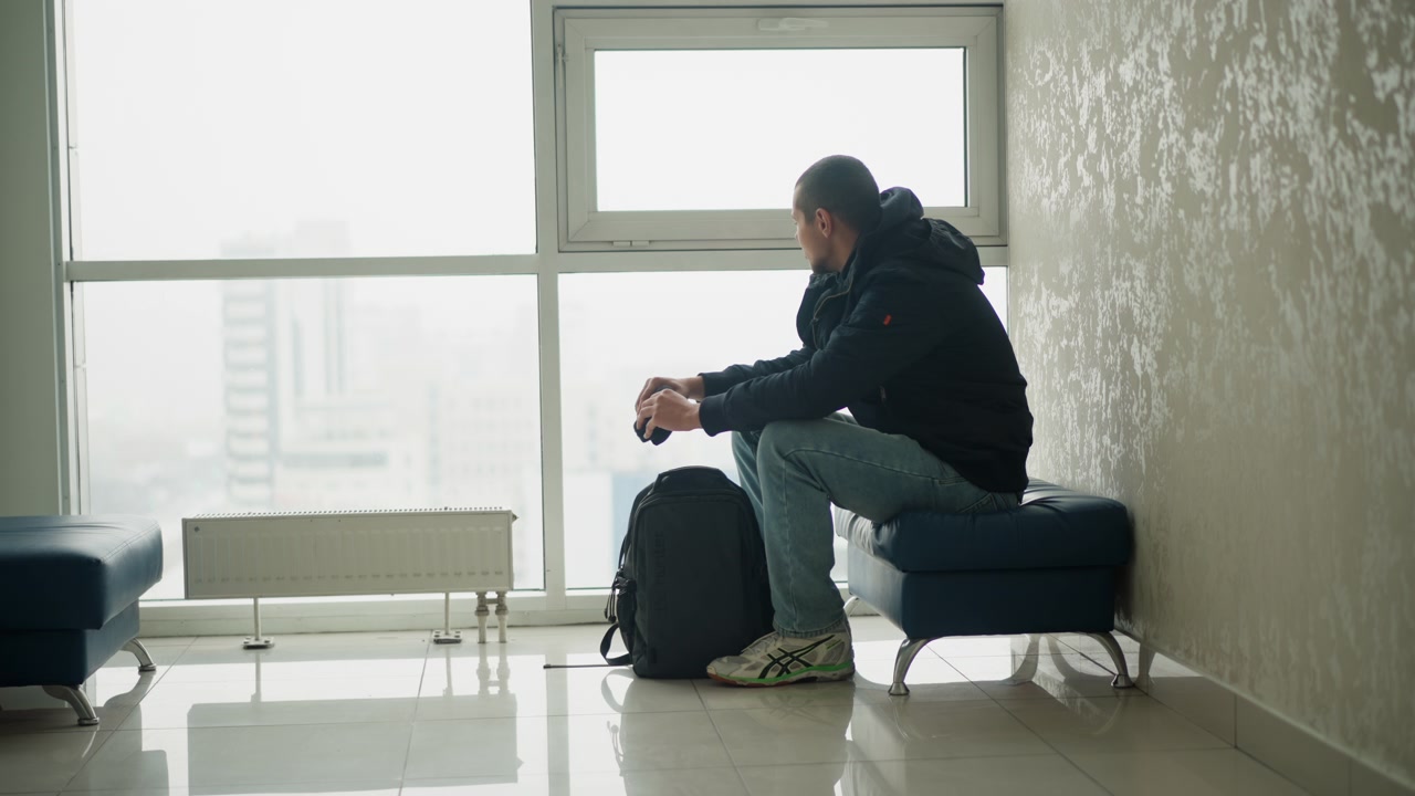 Man sits on blue bench in indoor waiting area holding small objects with minimal movement. Man wears casual clothing and has luggage beside him as man looks out large windows showing city