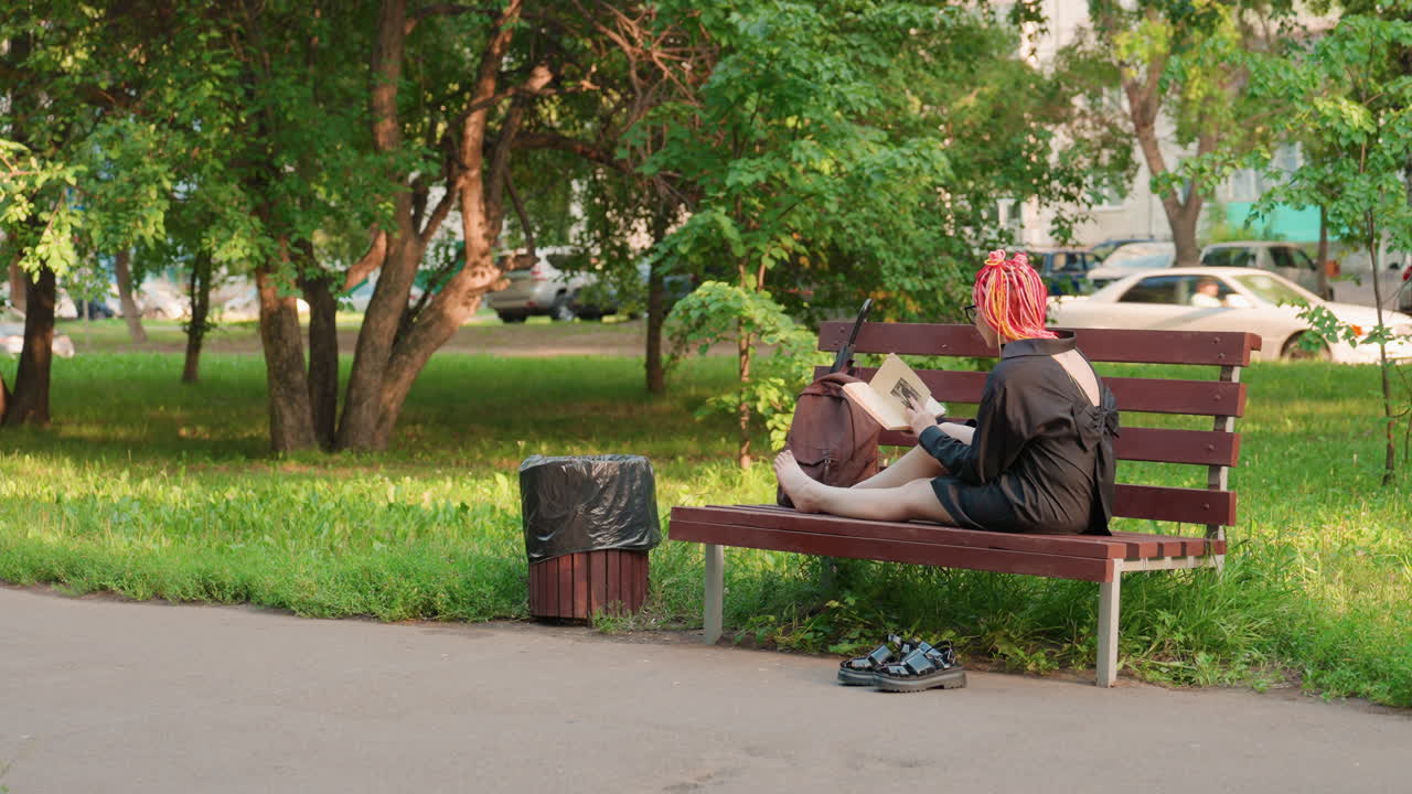 mujer sentada sola, mujer solitaria disfrutando del paisaje del parque al mediodía, joven relajándose sola en un banco del parque al mediodía, mujer absorta en un libro mientras está sentada en un banco del parque entre árboles y pasarelas