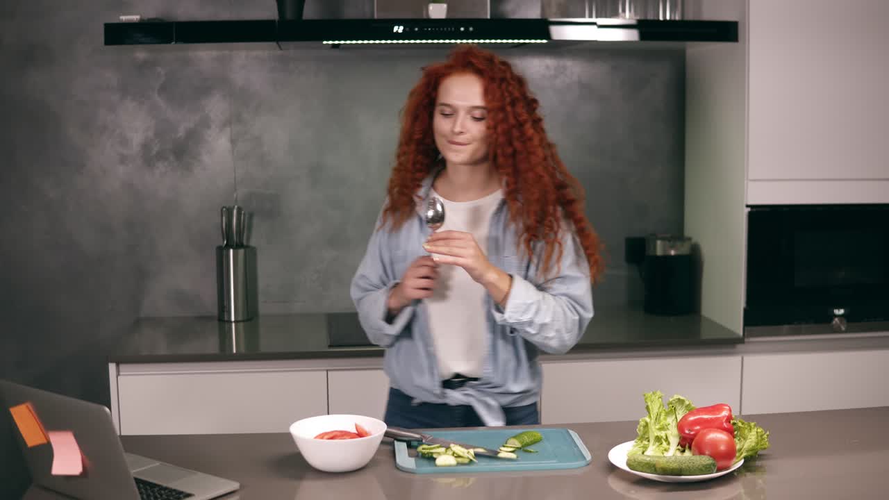 счастливая активная рыжая молодая женщина с ложкой в качестве микрофона поет песни и танцует во время приготовления пищи на кухне, беззаботно