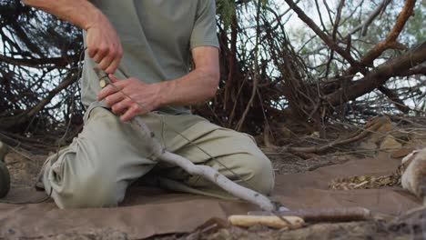 Caucasian-male-survivalist-tying-cord-to-branch-to-make-a-fire-bow-at-camp-in-wilderness