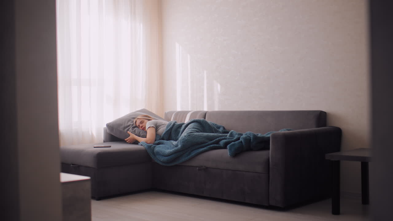 Student sleeping on couch adjusting position holding pillow wrapped in blanket phone by side as sunlight filters through curtains calm serene indoor atmosphere capturing relaxation comfort
