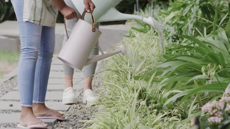 happy african american mother and daughter watering plants in garden, slow motion