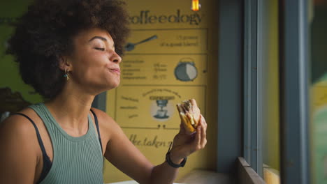 Cliente-Femenino-En-La-Ventana-De-La-Cafetería-Comiendo-Sándwich-De-Bagel-Para-El-Almuerzo