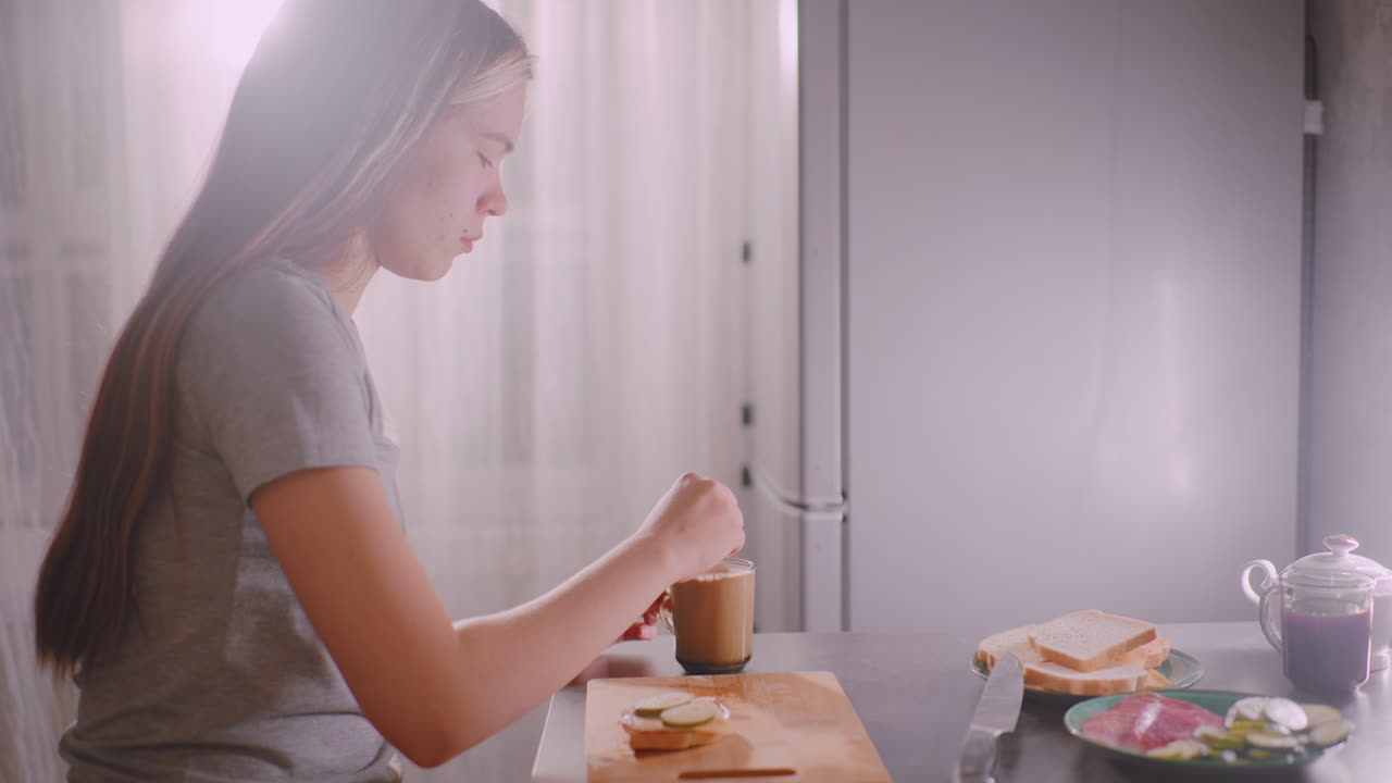 Girl stirring coffee at kitchen table with sunlight shining through curtain, sandwich ingredients and bread slices placed on cutting board beside her creating cozy breakfast atmosphere