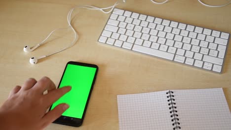 Hermosa-Mano-Femenina-Desplazándose-Y-Tocando-En-El-Teléfono-Con-Pantalla-Verde-En-Un-Lugar-De-Trabajo-Mínimo-Con-Teclado,-Auriculares-Y-Portátil