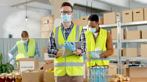 front view of caucasian male volunteer in facial mask checking donation list and smiling to the camera in charity warehouse