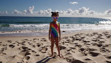 young girl stands confidently on a sandy beach, wearing a vibrant rainbow swimsuit and star shaped sunglasses, enjoying a sunny day by the ocean